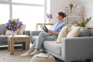 Oturma odasında kanepede ve vazoda leylak çiçekleri olan dergi okuyan genç bir kadın.
