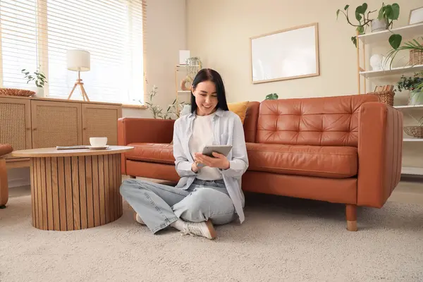 Genç bir kadın oturma odasındaki kahverengi kanepenin yanındaki halıda tablet bilgisayar kullanıyor.