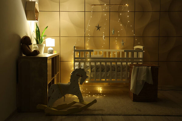 Интерьер детской спальни с детской кроваткой, комодом и светящимся светильником возле плитки стены ночью