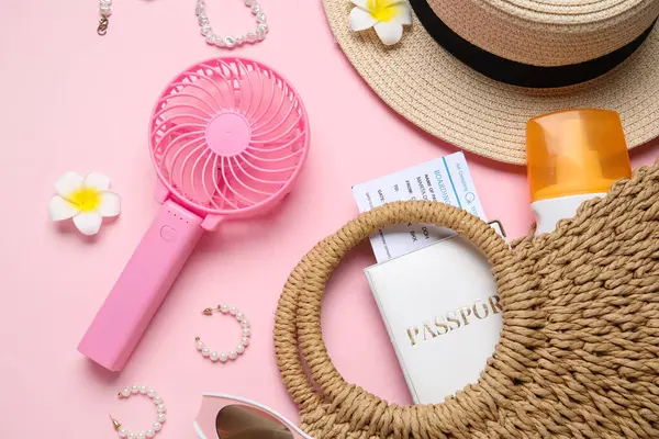 İçinde kadın aksesuarları, pasaport, bilet ve pembe arka planda mini fan olan bir çanta.