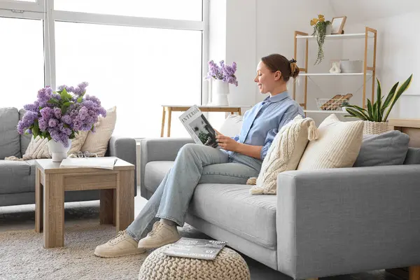 Oturma odasında kanepede ve vazoda leylak çiçekleri olan dergi okuyan genç bir kadın.