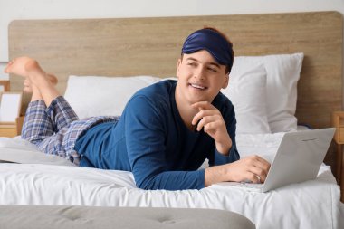 Güzel pijamalı, dizüstü bilgisayarı evde yatağında uzanmış yakışıklı genç bir adam.