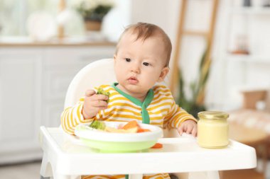 Evdeki yüksek sandalyede brokoli yiyen şirin bir bebek.
