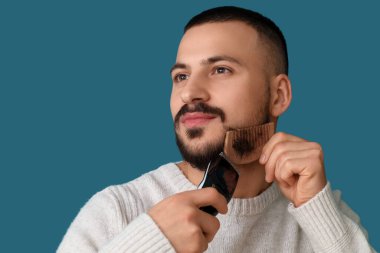 Tıraş makinesi ve mavi arka planda tarağı olan yakışıklı, sakallı bir adam.