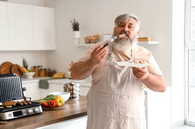 Mutfak masasındaki modern elektrikli ızgaranın üstünde pişmiş lezzetli sosislerle dolu tabağı olan olgun bir adam.