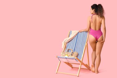 Hasır çantalı güzel bir Afro-Amerikalı kadın, pembe arka planda bir şişe güneş kremi ve iskemle.