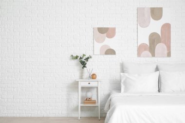 Güzel yatak odasının içi, rahat yatak odası, komodin, okaliptüs vazosu ve beyaz tuğla duvarda resimler.