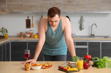 Mutfaktaki fast food ve sağlıklı yemek arasında seçim yapan üzgün, şişman genç bir adam. Kilo alma kavramı