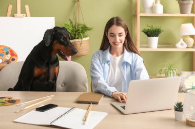 Güzel Doberman köpeği paletin yanında ve güzel, mutlu bir kadın evde dizüstü bilgisayarla çalışıyor.