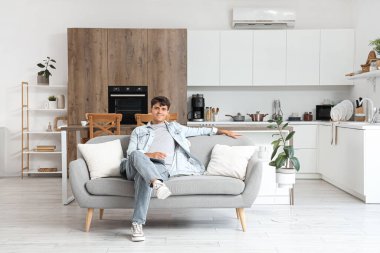 Mutfaktaki kanepede oturan klimalı uzaktan kumandalı yakışıklı genç adam.