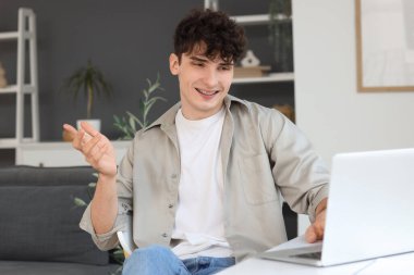 Genç adam evdeki masada dizüstü bilgisayarla sohbet ediyor.