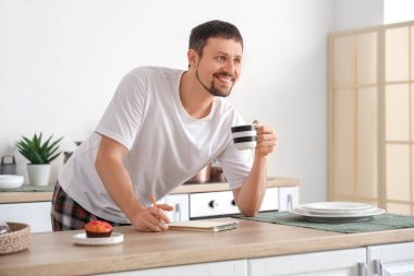 Evde pijamalı, defterli, kahveli ve kekli genç adam.