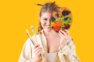 Sarı arka planda boya fırçası ve paleti olan kadın sanatçı.