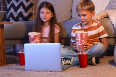 Küçük çocuklar ellerinde patlamış mısırla evde dizüstü bilgisayar seyrediyorlar.