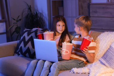 Küçük çocuklar ellerinde patlamış mısır ve sodayla evde dizüstü bilgisayar seyrediyorlar.