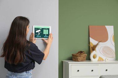 Genç bir kadın odasının duvarında, arka tarafında akıllı ev güvenlik kontrol paneli kullanıyor.