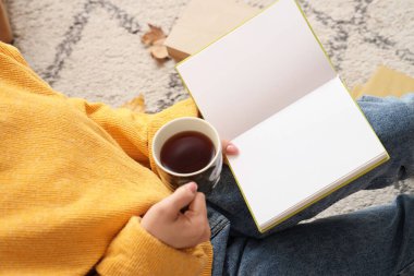 Elinde kahve fincanı olan kadın evde halıda kitap okuyor, yakın plan.