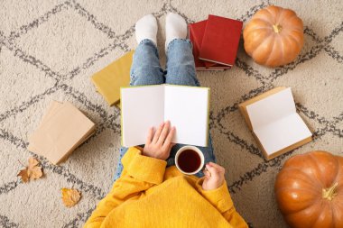 Kahve fincanı ve balkabağı olan bir kadın halıda kitap okuyor.