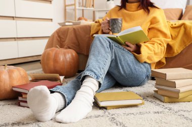 Kahve fincanı ve balkabağı olan bir kadın evde halıda kitap okuyor.