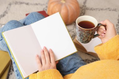 Elinde kahve fincanı olan kadın evde halıda kitap okuyor, yakın plan.