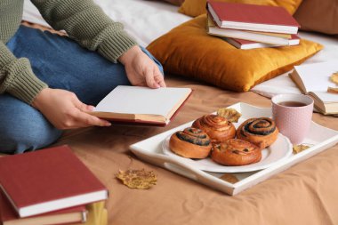 Evde, yatakta çörekleri ve sonbahar yapraklarıyla kitap okuyan bir kadın.