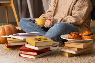 Elinde çay fincanı, kitapları ve çörekleri olan bir kadın evdeki halıda oturuyor, yakın plan.
