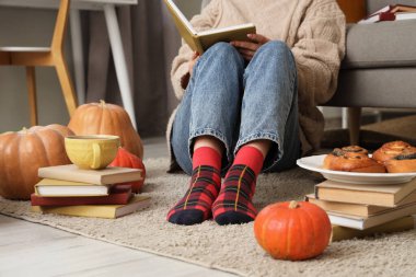 Evde halıda balkabağı ve çöreklerle kitap okuyan bir kadın.