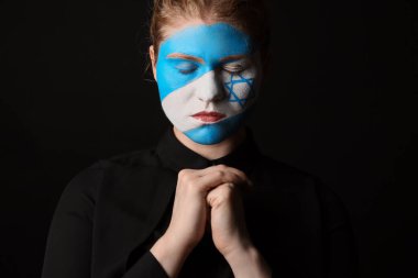 Yüzünde İsrail bayrağı olan dua eden genç bir kadın siyah arka planda, yakın plan. Uluslararası Soykırım Anma Günü