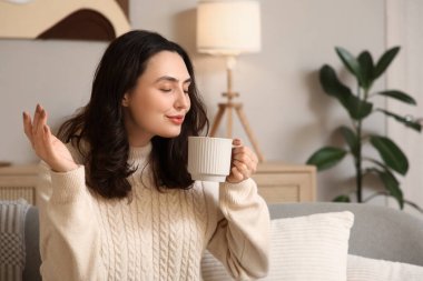 Evdeki kanepede oturan bir fincan sıcak çikolatalı güzel, genç ve mutlu kadın.