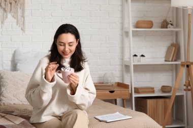 Güzel, genç, mutlu bir kadın. Bir fincan sıcak çikolata ve marşmelovla evde yatakta oturuyor.