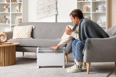 Evde elektrikli konteyner ısıtıcısının yanındaki koltukta oturan çaylı genç adam.