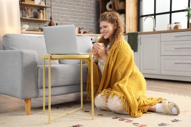Mutfakta dizüstü bilgisayar kullanan, beyaz kahve ve battaniyeli güzel genç bir kadın.