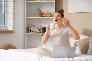 Bir fincan kahve ve dizüstü bilgisayarlı güzel bir kadın yatak odasında oturuyor.