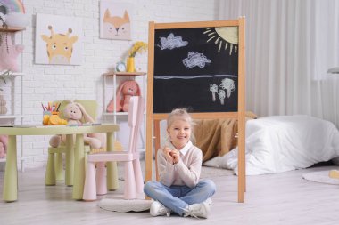 Yatak odasındaki karatahtanın yanında oturan tatlı küçük kız.