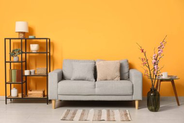 Konforlu kanepe, raf ünitesi ve sarı duvarın yanında çiçek açan ağaç dalları.