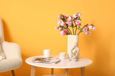 Koltuk ve kahve masası. Odada sarı duvarın yanında çiçek açan manolya dalları var.