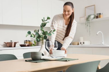 Genç bir kadın mutfaktaki yemek masasına temiz tabaklar koyuyor.