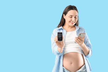 Mavi arka planda glukometresi ve cep telefonu olan hamile bir kadın. Gebelik Diyabet Konsepti