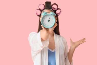 Saç bigudileri ve pembe arka planda çalar saati olan geç kalmış genç bir kadın. Yakın plan.