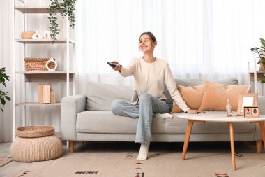 Oturma odasındaki kanepede uzaktan kumandayla televizyon izleyen genç bir kadın.