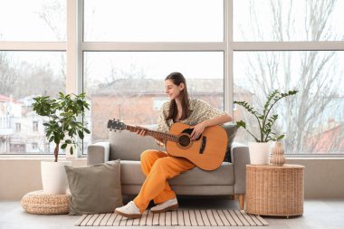 Evdeki kanepede gitar çalan genç bir kadın.