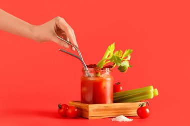 Saman ve kırmızı arka planda pastırmalı Bloody Mary kokteyli kavanozu olan bir kadın eli.