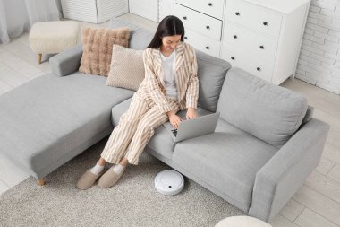 Pijamalı genç bir kadın evinde robot elektrikli süpürgeyle kanepede dizüstü bilgisayar kullanıyor.