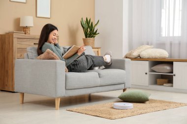 Evde elektrikli süpürgeyle kanepede kitap okuyan genç bir kadın.