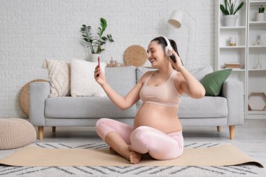 Evde yoga minderinde cep telefonu kullanan kulaklık takan hamile bir kadın.