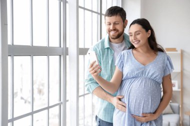 Mutlu hamile çift evdeki pencerenin yanında cep telefonu kullanıyor.