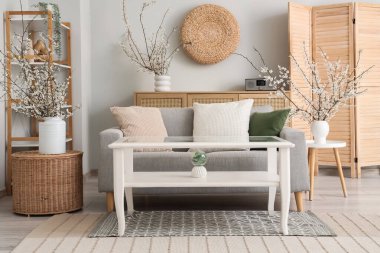 Aydınlık oturma odasının içinde, kanepe ve vazolarda çiçek açan dallar var.