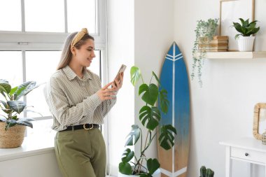 Yatak odasında sörf tahtası olan cep telefonu kullanan genç bir kadın.