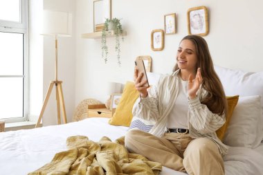 Cep telefonlu genç bir kadın yatak odasında görüntülü sohbet ediyor.