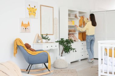 Bebek kıyafetleri asan genç bir kadın çocuk odasında, arkadan bakıyor.